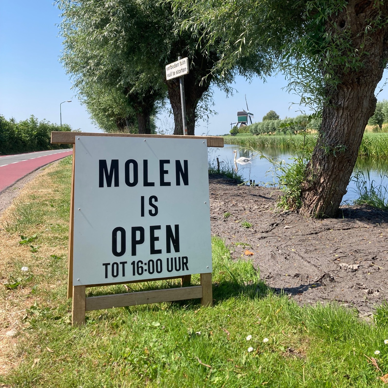 Foto met op de voorgrond een wit klapbord met daarop geschreven 'Molen is open tot 16:00 uur'. Op de achtergrond zie je tussen de knotwilgen door een zwaan zwemmen, en de kockengense molen.