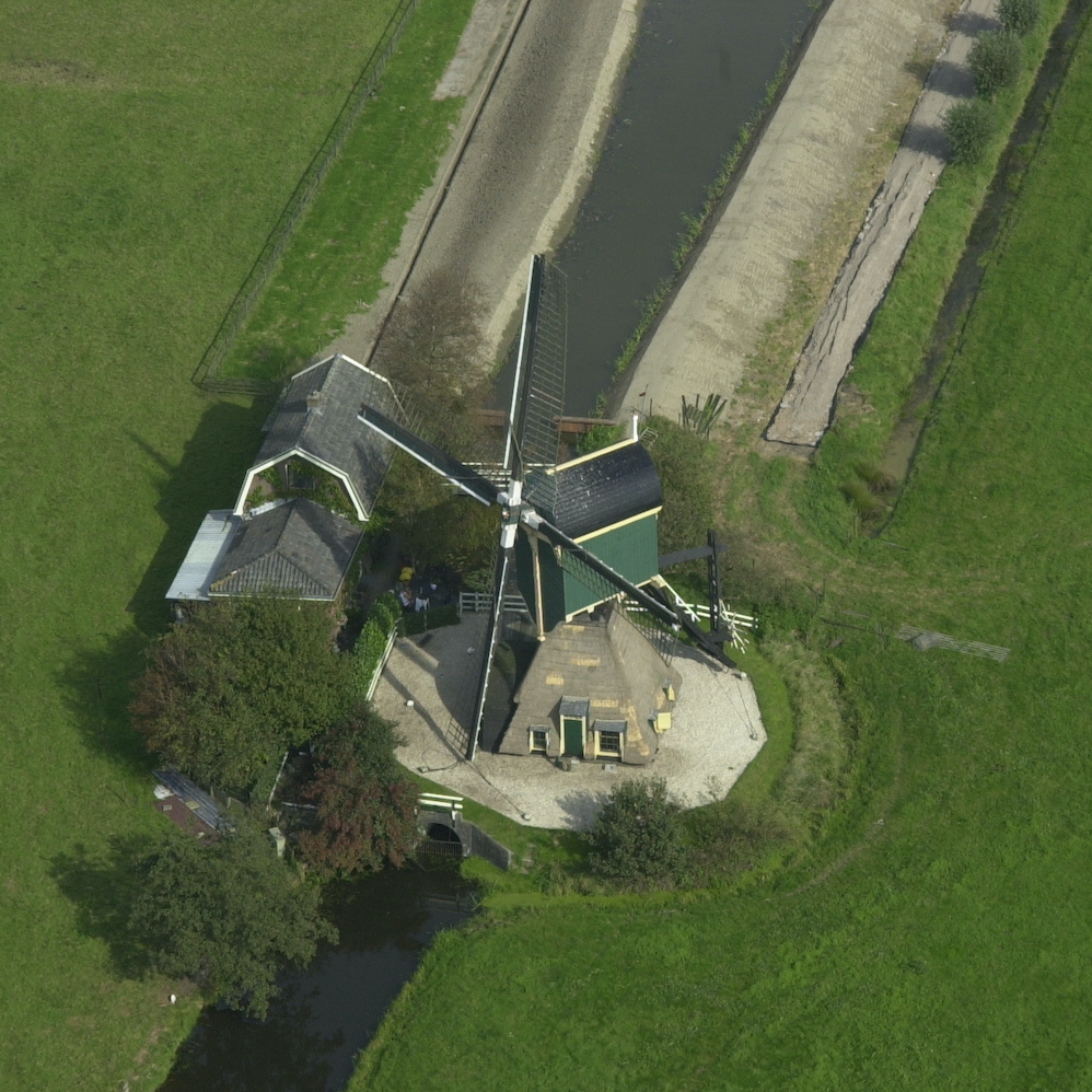 Een foto van de Kockengense Molen vanuit een vogelperspectief vanuit de lucht. Je ziet ook het woonhuis naast de molen, een stukje van de voorwaterloop en de achterwaterloop, de weg naar de molen met knotwilgen er naast, en de weilanden naast de molen.